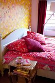 Rote Bettwäsche mit floralem Muster auf Doppelbett mit weißem Kopfteil, vor tapezierter Wand mit Blumenmuster