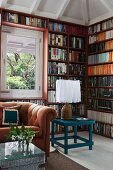 Seating area with caramel-coloured sofa, wicker trunk and petrol blue table in front of full-wall bookcases