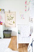 Vintage-style botanical illustration on wall above black and white patterned basket on floor next to white cot in nursery