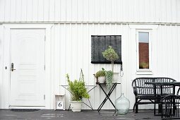 House with white-painted wooden façade, potted plants on table and floor and black outdoor furniture on terrace