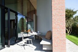 Sonnenbeschienene, möblierte Terrasse mit gebogener Beton Bank an Wand, gegenüber verglaste Fassade zeitgenössischer Architektur