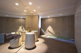 Zylindrisch geformte Standwaschbecken mit Messing Standarmatur vor verspiegelter Wand in luxuriösem Bad, im Hintergrund in Boden eingelassene Badewanne
