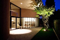 Spacious paved terrace outside contemporary house with curved glass façade and illuminated interior at twilight