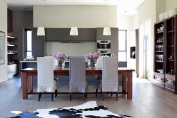 Chairs with loose covers in different shades of grey around long dining table, open-plan fitted kitchen in background, colonial-style wooden furniture and animal-skin rug