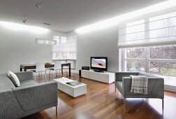 Pale grey, elegant sofa set around low white table opposite TV on low sideboard on elegant walnut parquet floor in modern interior with half-closed roller blinds on French windows