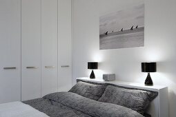 Double bed with grey patterned bed linen and black table lamps on headboard shelf in modern bedroom with white fitted wardrobes