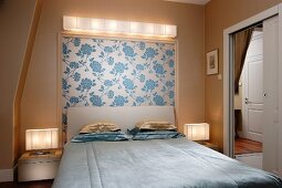 Schlichtes Doppelbett mit Polster Kopfteil und Kissenstapel vor Wand mit Blumentapete als Bild gerahmt, darüber Beleuchtung, seitlich offene Tür und Blick in Nebenzimmer
