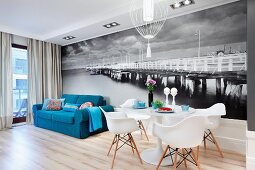 Tulip table, Plastic Armchairs and blue sofa in front of black and white mural wallpaper depicting old pier in the Baltic Sea