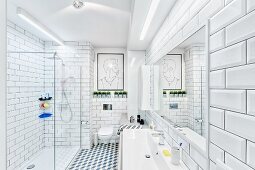 White, modern bathroom with tiled walls & floor, strip lights over washstand & glazed, floor-level shower; modern artwork above wall-mounted toilet