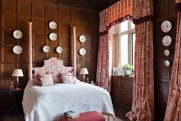 Toile-de-jouy curtains and double bed with ornate headboard in wood-panelled bedroom with decorative china plates on walls