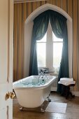 Free-standing bathtub in front of window with Tudor arch and draped petrol-blue curtains combined with multicoloured striped wallpaper