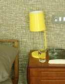 Yellow, retro bedside lamp on wooden bedside cabinet against wallpapered wall