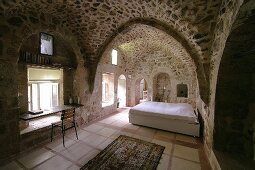 Historisches Wohnhaus mit Mauer und Gewölben aus Kalkstein, im Hintergrund grosses Bett in offenem Schlafbereich, seitlich improvisierter Arbeitsplatz vor Fenster