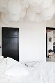 Doppelbett mit weisser Bettwäsche unter einem Meer von Papierlampen an Decke, im Hintergrund schwarz lackierte Zimmertür