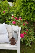 Jugs and cushions on white-painted bench in front of flowering rhododendron
