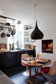 Essplatz mit weißen Klassikerstühlen unter schwarzer Pendelleuchte vor Küchenzeile in eklektischem Ambiente mit offenem Kaminfeuer und Grilleinsatz