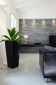 Black planter on floor, partially visible leather couch, illuminated stone wall and low, floating black sideboard