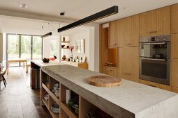 Lange Küchentheke aus Beton unter stabförmigen Hängeleuchten, gegenüber Einbauschrank mit Holzfront