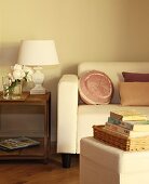 Bücherstapel auf Hocker gegenüber Sofa mit eleganten Kissen, daneben Beistelltisch aus Holz und Tischlampe mit weißem Schirm im Vintage Stil