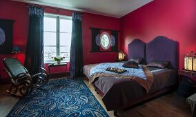 Double bed with purple headboard, rocking chair and paisley rug in bedroom with red walls