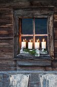 Rustikaler Adventskranz mit vier brennenden Kerzen im Fenster eines verwitterten Holzhauses