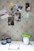 Postcards stuck on elegant fabric-covered pinboard above pen holders and vase of flowers on white desk