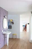 Pedestal sink and mirror on wall with retro wallpaper in open-plan interior with view of woman through multiple open doorways