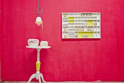 Übertöpfe und runder Beistelltisch mit neonfarbenen Kordeln umwikelt, aufgehängter weisser Setzkasten mit gelben Farbakzenten an Wand in Pink