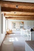 White dining area with rustic wood-beamed ceiling, artworks and pattern of light and shade on concrete floor