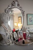 Family photos, perfume bottles and bust used as jewellery stand in front of ornate silver mirror on top of chest of drawers