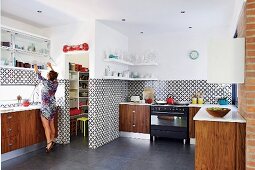 Wooden base units against black and white, geometric wall tiles and woman opening wall cabinet in open-plan kitchen