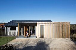 Blue sky above modern house with family in open front door and vertical wooden slat structure covering facade in rural area