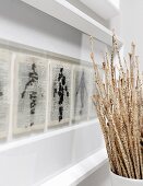 Dried stems in vase next to paintings on printed pages in white picture frame