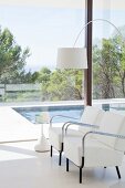 Two elegant white armchairs, arc lamp and side table in front of glass wall with view of pool