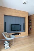 Designer armchair in front of wooden living room wall with flatscreen TV and speakers in niche in open-plan living room