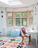 Brightly patterned rug in front of comfortable beanbag in window bay and antique chair at white table to one side in teenager's bedroom