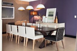 White leather chairs at long, dark-wood table below pendant lamps with white and gold lampshades; purple-painted accent wall in background