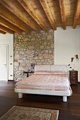 Weisses, modernes Bett mit elegantem Überwurf im Schlafzimmer mit Holzbalkendecke und Natursteinwand-Ausschnitt