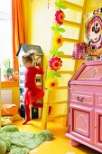 Pinkfarbener Sekretär in Kinderzimmer mit gelber Wand, Boden und Leitertreppe, davor Mädchen