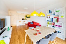 Essplatz mit gelben Pendelleuchten vor Bücherregal, dahinter Medienboard, rotes Sofa und offene Küche mit Frühstückstresen