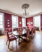 Chairs with patterned upholstery around polished wooden table below chandelier in grand dining room with red walls