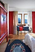 Schlafraum in Rot- und Blautönen mit lackiertem Ethno-Schrank und kunsthandwerklicher Sitzbank, verschiedene Vorhänge vor den Fenstern - in einem Fertighaus