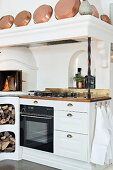 White, country-house-style kitchen counter with integrated oven, fireplace, firewood store and copper pans on mantel hood