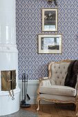 Rococo armchair with gilt frame next to rounded, tiled corner fireplace and against blue-patterned wallpaper