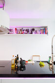 Kücheninsel mit schwarzer, polierter Platte, darauf Küchenutensilien, gegenüber hellviolett beleuchtetes Wandregal
