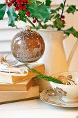 Weihnachtskugel mit Waldmotiv an Stechpalmenzweigen in einer Kanne, davor Bücherstapel und Tasse