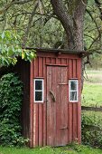 Old, wooden toilet outhouse painted Falu red hidden between bush and tree in garden
