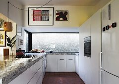Moderne Einbauküche in Weiß, auf Unterschränken massive Steinplatte, im Hintergrund breites Fenster mit Blick auf Mauer