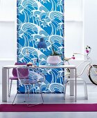 Schlichter weisser Esstisch mit Hocker und Stühlen vor blau-weisser Stoffbahn mit stilisiertem Wellenmotiv