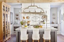 Bar stools with loose covers at island counter below kitchen utensils hung from pendant lamp in country-house kitchen